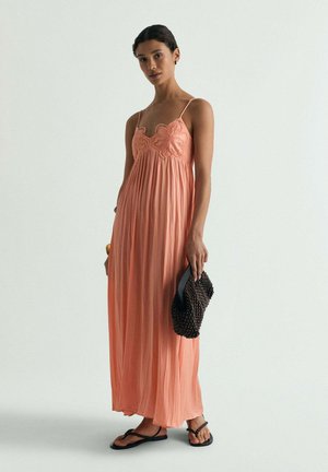 Mujer con un vestido largo coral plisado con bordado floral, llevando un bolso oscuro tejido y sandalias negras.