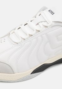 Chaussure de sport blanche avec tige en cuir lisse, motifs ondulés cousus, semelle en caoutchouc et bout en daim gris. Comprend des lacets blancs et un logo.