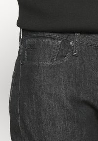 Mörka jeans i denim med synlig framficka, kontrasterande sömmar och en diskret logga på midjebandet. Texturerat tyg med en mjuk yta.