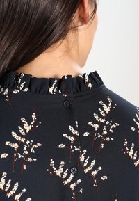 Žena ze zády s tmavohnědými vlasy, oblečená v černé blůze s bílými květinovými vzory, volánkovým límcem a třemi černými knoflíky na výstřihu.