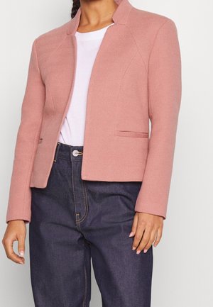 Vrouw die een gestructureerd roze colbert draagt over een wit shirt, gecombineerd met donkerblauwe high-waisted jeans, staand tegen een effen achtergrond.