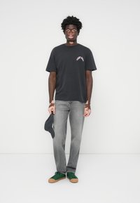 Sort bomulds t-shirt med trykt grafik, parret med grå denimjeans og grønne sneakers. Holder en mørk baseballkasket.