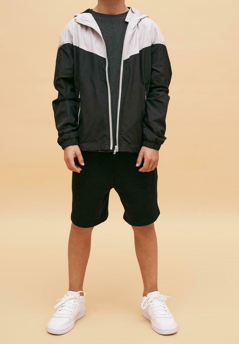 Personne portant une veste zippée noire et blanche, un short foncé et des baskets blanches, debout devant un fond beige avec les mains détendues sur les côtés.
