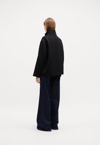 Čierna oversized bunda s vysokým golierom, vyrobená z hladkej látky, v kombinácii s širokými tmavými denimovými nohavicami. Jednoduchý a štruktúrovaný dizajn.