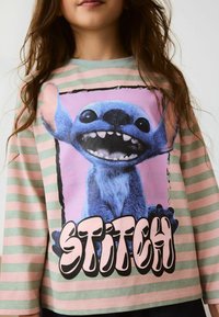 Long-sleeve shirt with pastel pink and green stripes. Features a graphic of a blue character with large ears and the text "STITCH" in bold.