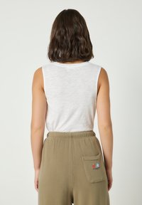 Žena s krátkými vlnitými hnědými vlasy, oblečená v bílém tílku bez rukávů a olivově zelených kalhotách s zadní kapsou zdobenou malou výšivkou ovoce.