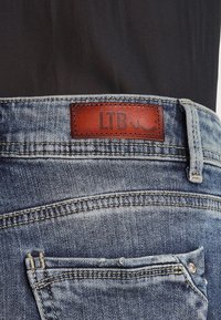 Gros plan sur la poche arrière d'un jean en denim bleu et sur l'étiquette de marque en cuir marron portant le texte "LTB", portée avec un haut noir.