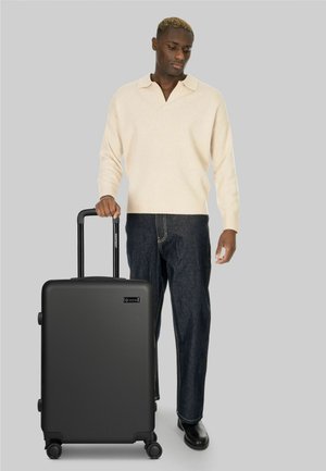 Hombre con suéter beige y jeans oscuros de pie con una maleta negra con ruedas contra un fondo gris liso.