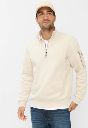 Mand iført beige hættejakke med lynlås og lomme på ærmet, blå jeans, beige kasket, der ser let til siden mod en ensfarvet baggrund.