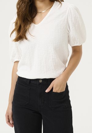 Femme portant un chemisier blanc texturé à manches courtes et un jean noir taille haute, une main dans la poche, sur fond uni.