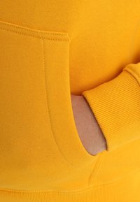 Tissu de sweat-shirt jaune à texture côtelée, avec une poche avant et des coutures latérales. Gros plan sur le tissu et les détails de couture.