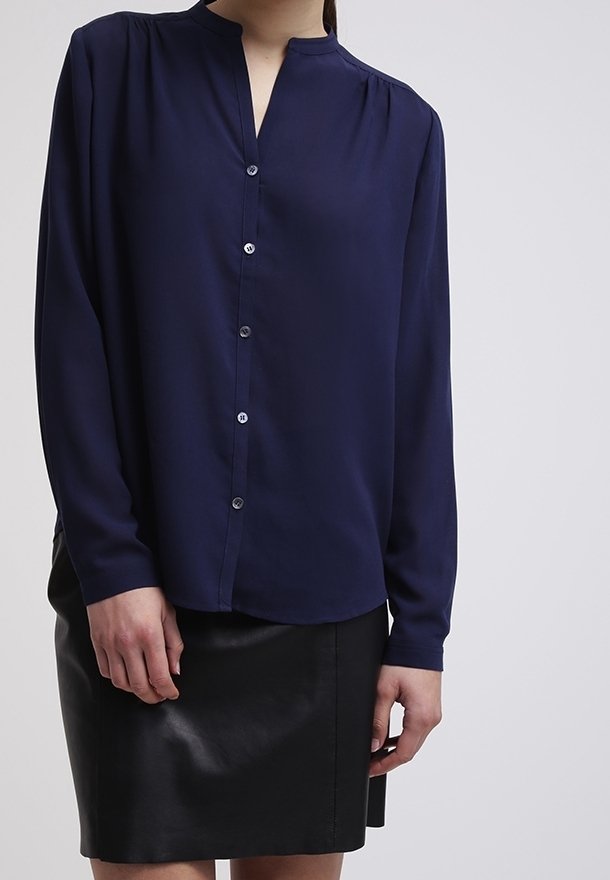 Blusa lunga blu navy con scollatura a V, frontale con bottoni e tessuto morbido e fluido. Indossata con una gonna in pelle nera attillata.