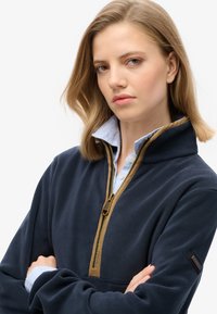Marineblauwe fleece pullover met een hoge kraag, gouden rits en bruine accenten. Gelaagd over een lichtblauw overhemd met strepen.