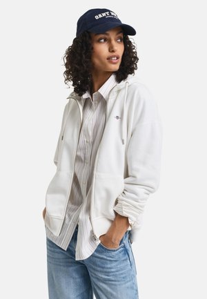 Felpa bianca con zip e coulisse, indossata sopra una camicia a righe chiara con bottoni. Jeans in denim blu e un cappellino da baseball blu navy con testo bianco.