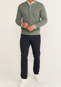 Gestricktes, weiches grünes Poloshirt mit Kragen und drei Knöpfen, kombiniert mit marineblauen Hosen und weißen Turnschuhen. Glatte Textur, lässige Passform.