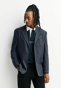 Marineblauer Fischgrätenblazer mit einer einzigen Brusttasche, kombiniert mit einem dunklen Poloshirt. Der Stoff wirkt strukturiert und maßgeschneidert.