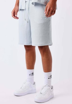 Gambe maschili che indossano pantaloncini grigio chiaro testurizzati, sneakers bianche e calzini bianchi con loghi neri su sfondo uniforme.