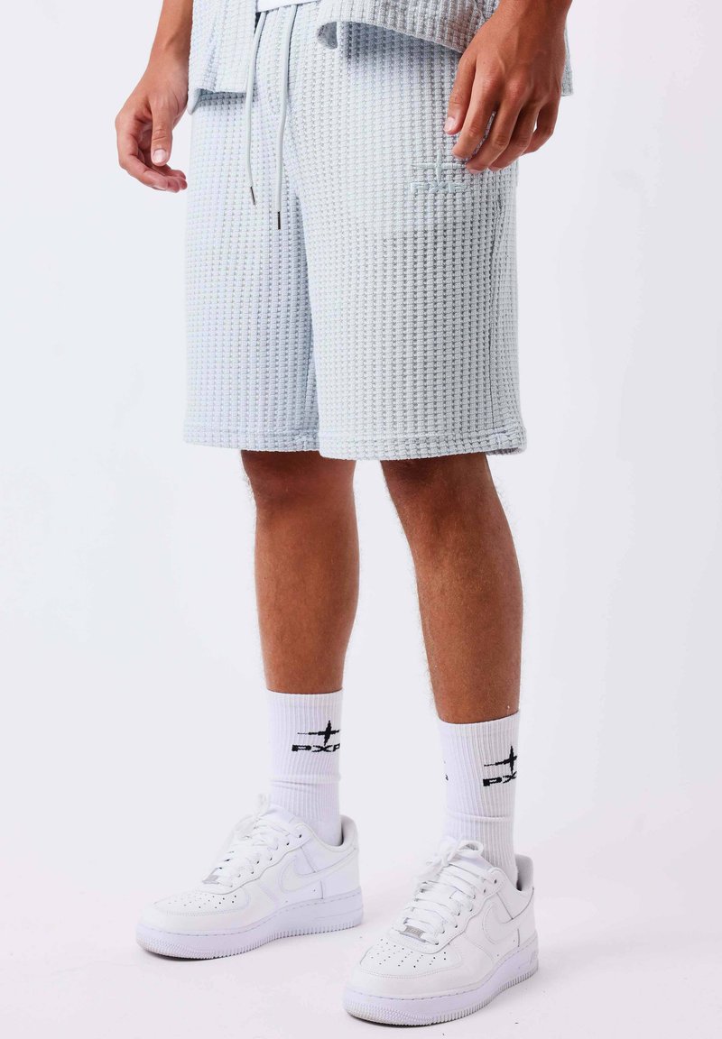Jambes masculines portant un short texturé gris clair, des baskets blanches et des chaussettes blanches montantes avec des logos noirs sur un fond uni.