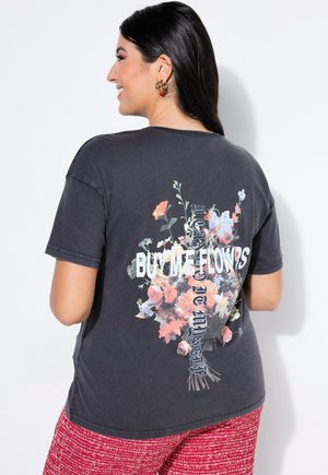 Pitkät tummaat hiukset omaava hymyilevä nainen, jolla on yllä tummanharmaa T-paita, jossa on värikäs kukkakimppu ja teksti "OSTA MINULLE KUKKIA" takana.