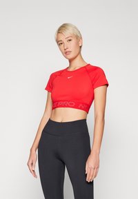 Top sportiv scurt roșu, cu mâneci scurte și o bandă țesută care prezintă logo-ul. Combinație cu leggings negri, cu talie înaltă. Textură netedă.