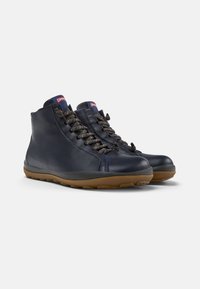Zapatos altos de cuero azul marino con superficie texturada, punta redondeada y suela de goma marrón en contraste. Presenta un cierre de cordones oscuros y acentos de logotipo.