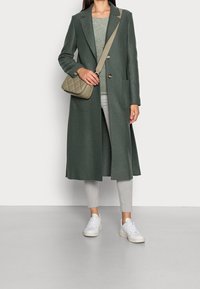 Pitkä vihreä takki pehmeästä kankaasta, yhdistettynä harmaaseen yläosaan, vaaleisiin harmaisiin farkkuihin ja valkoisiin lenkkareihin. Mukana myös vahva beige olkalaukku.