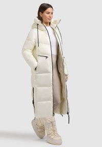 Lang, cremefarvet dynejakke med hætte, der har sorte lynlåsdetaljer og lommer. Glat tekstur med quiltede sektioner for isolering.