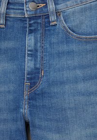 Jean en denim bleu en gros plan montrant la poche avant, les passants de ceinture, les coutures et la fermeture par bouton métallique.