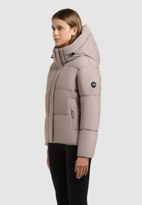 Hellbeige Steppjacke mit großer Kapuze, funktionalem Design, Reißverschlusstaschen und Logoaufnäher am Ärmel. Glatte, gepolsterte Textur.