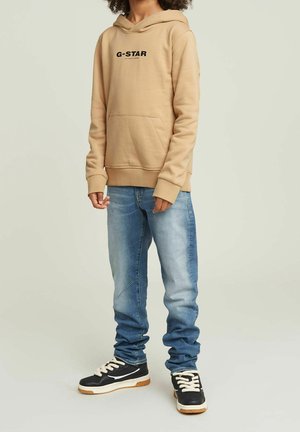 Beżowa bluza z kapturem G-Star z czarnym logo, kieszenią kangurka i ściągaczami przy mankietach. W połączeniu z jasnoniebieskimi dżinsami i czarnymi trampkami z białymi sznurowadłami.