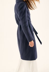 Tmavě modrý kabát z vlněné směsi s texturovaným povrchem, dlouhými rukávy, páskem v pase a širokým límcem. Střih na míru s bočními kapsami.