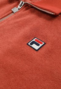 Nærbillede af et rødt tekstureret lynlåsbeklædningsstykke med en sølvfarvet lynlås og et lille broderet logo med røde, marineblå og hvide elementer.