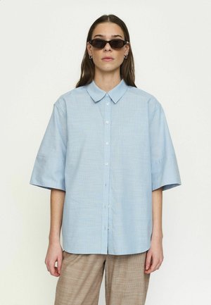 Nő, aki túlméretezett világoskék gombos inget, barna kockás nadrágot és keskeny fekete napszemüveget visel, egyszínű háttér előtt áll.