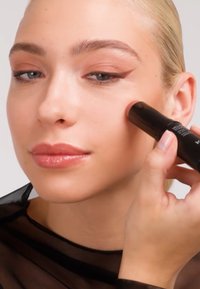 Kremrouge i stift med matt svart tube, påført på kinnet. Modellen har glatt hud, nøytral sminke og lyse hår.