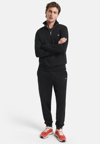 Felpa nera con zip e collo alto, abbinata a pantaloni jogger neri e scarpe sportive rosse. Presenta un logo sul petto e polsini elasticizzati.