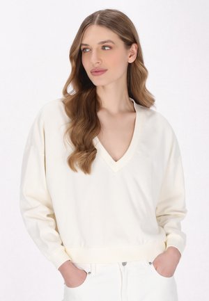 Maglione crema a V con maniche lunghe e polsini a coste, realizzato in tessuto morbido, abbinato a pantaloni bianchi. Design minimalista.