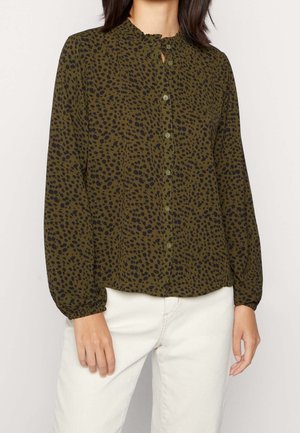 Kobieta w oliwkowo-zielonej bluzce z długim rękawem z czarnymi cętkami w panterkę i białych spodniach, stojąca na jednolitym tle.