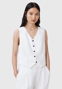 Weißes ärmelloses Oberteil mit V-Ausschnitt, ausgestattet mit einer sechsknöpfigen Frontverschluss, seitlichen Taschen und tailliertem Schnitt. Minimalistisches Design und glatte Textur.