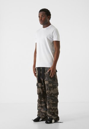 Weißes T-Shirt mit kurzen Ärmeln und Cargohosen im Camouflage-Muster mit mehreren Taschen, ausgestattet mit schwarzen Hardware-Details und einem lockeren Schnitt. Schwarze Sneakers.