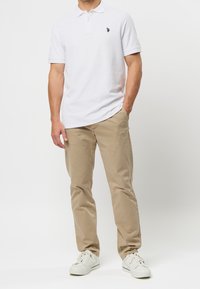 Bílá polo košile s tříknoflíkovým zapínáním a vyšitým logem, spárovaná s béžovými chinos kalhotami a bílými teniskami. Hladká textilie.