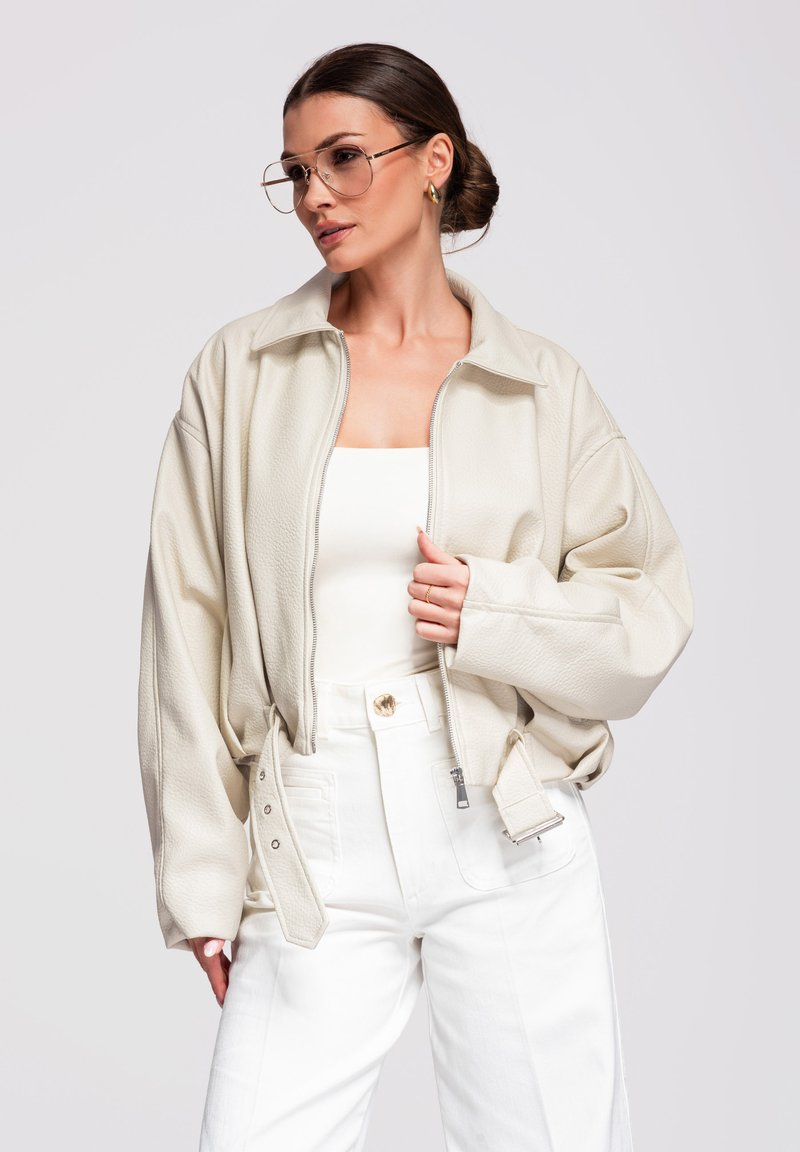 Mujer con chaqueta beige de gran tamaño, camiseta blanca, pantalones blancos de cintura alta, pendientes de aro dorados y gafas aviador grandes, posando frente a un fondo liso.