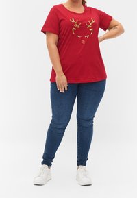 Rød t-shirt med korte ærmer, udsmykket med gyldne palietter i form af et hjortegevir og en næse på fronten; parret med blå skinny jeans og hvide sneakers.