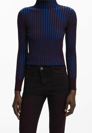 Osoba o ciemnej karnacji, ubrana w dopasowany, długi rękaw, fioletowy golf z pionowymi niebieskimi paskami oraz ciemne spodnie z wysokim stanem.