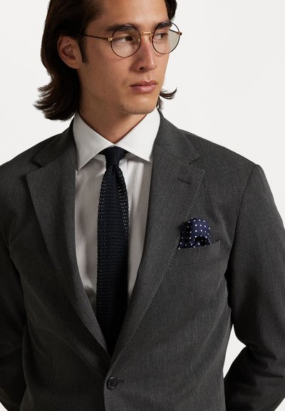 Grijze op maat gemaakte colbert met notch-lapels, gecombineerd met een witte overhemd en een gestructureerde donkere stropdas. Een marineblauwe pochet met witte stippen is zichtbaar.