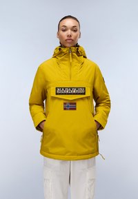 Gelbe wasserfeste Jacke mit hohem Kragen, Reißverschluss vorne und großer Brusttasche. Enthält ein Patch mit der norwegischen Flagge und ein Logo.