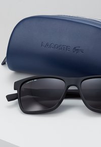 Ochelari de soare negri, cu finisaj mat și lentile întunecate, așezați în fața unei carcase de protecție albastre, inscripționate cu "LACOSTE."