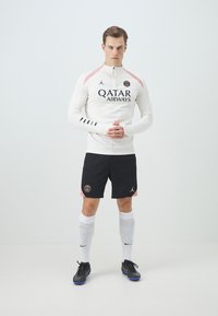 Vit långärmad träningsskjorta med rosa accenter, svarta shorts med en logotyp, vita strumpor och svarta fotbollsskor på en neutral bakgrund.