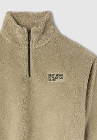 Beżowy polarowy pulower z czarnym półsuwnym zamkiem i naszywką "New York Athletics Club" na piersi.