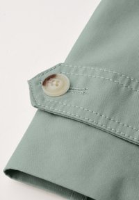 Zielony materiał, mankiet zapinany na guziki z jasnym guzikiem, delikatna struktura prążkowa, czyste krawędzie i kontrastujące detale przeszycia.
