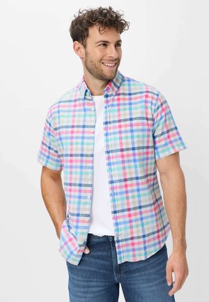 Mann mit lockigem Haar trägt ein kurzärmeliges Pastell-Karohemd über einem weißen T-Shirt und blauen Jeans, lächelt und schaut nach rechts.